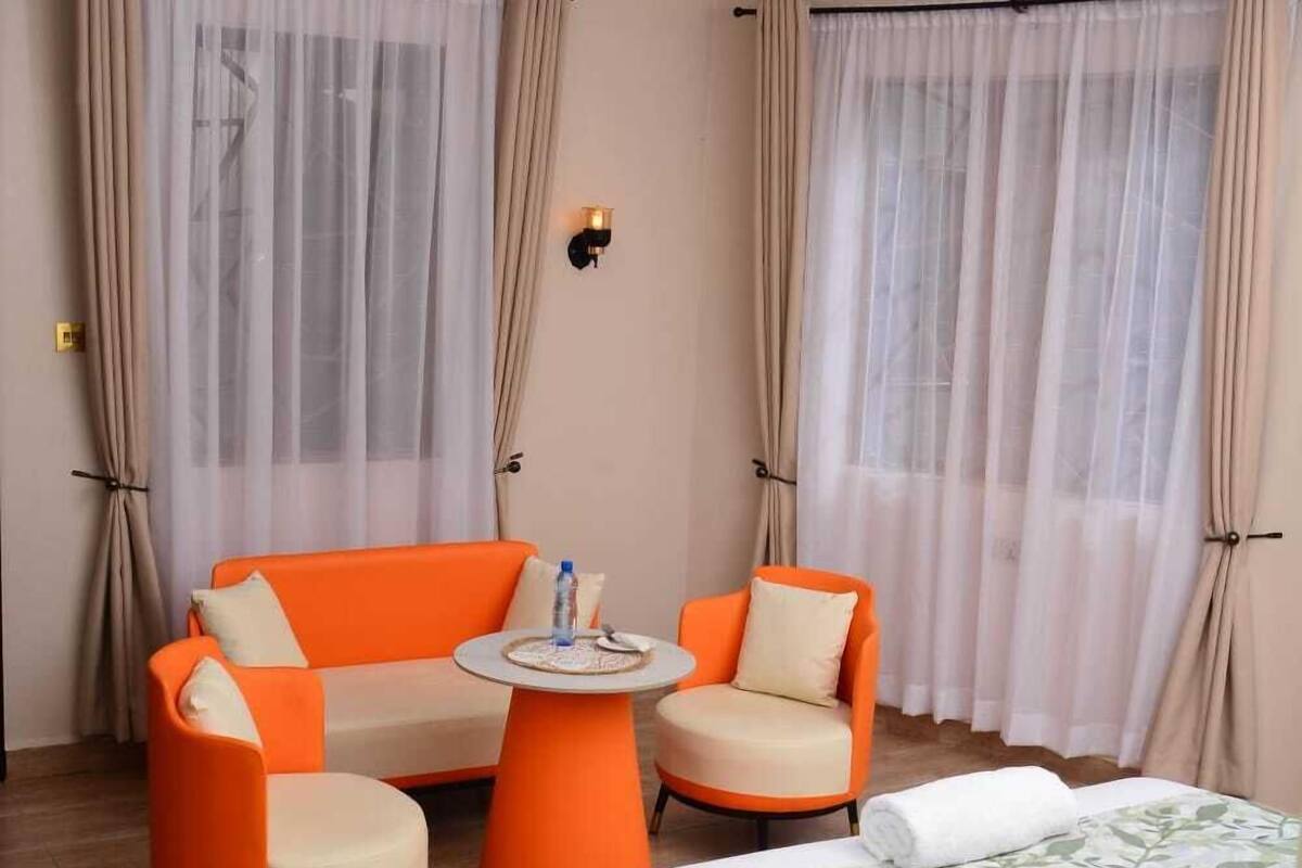 A hotel room with a bed, two orange armchairs, a small round table, and sheer curtains.