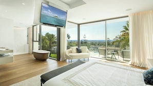 Ein modernes Schlafzimmer mit einem großen Bett, einem an der Wand montierten Fernseher, einem Balkon mit Meerblick und einer Sitzecke mit Sofa und Sesseln.