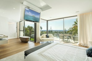 Ein modernes Schlafzimmer mit einem großen Bett, einem an der Wand montierten Fernseher, einem Balkon mit Meerblick und einer Sitzecke mit Sofa und Sesseln.