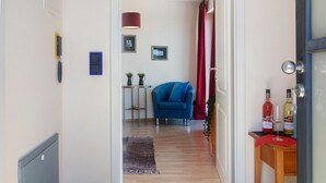 Une pièce avec une chaise bleue, une petite table sur laquelle se trouve une plante, un rideau rouge et deux bouteilles de vin posées sur une table d’appoint en bois.