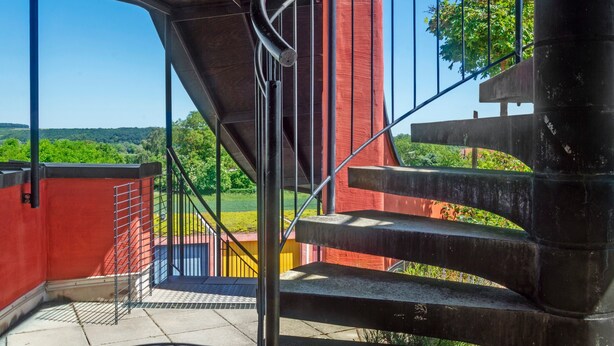 Un escalier avec des rampes en métal menant à un espace extérieur offrant une vue sur la verdure et un ciel dégagé.