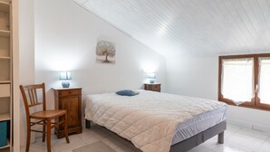 A bedroom with a bed, two bedside lamps, a wooden chair, a small cabinet, and a window with curtains.