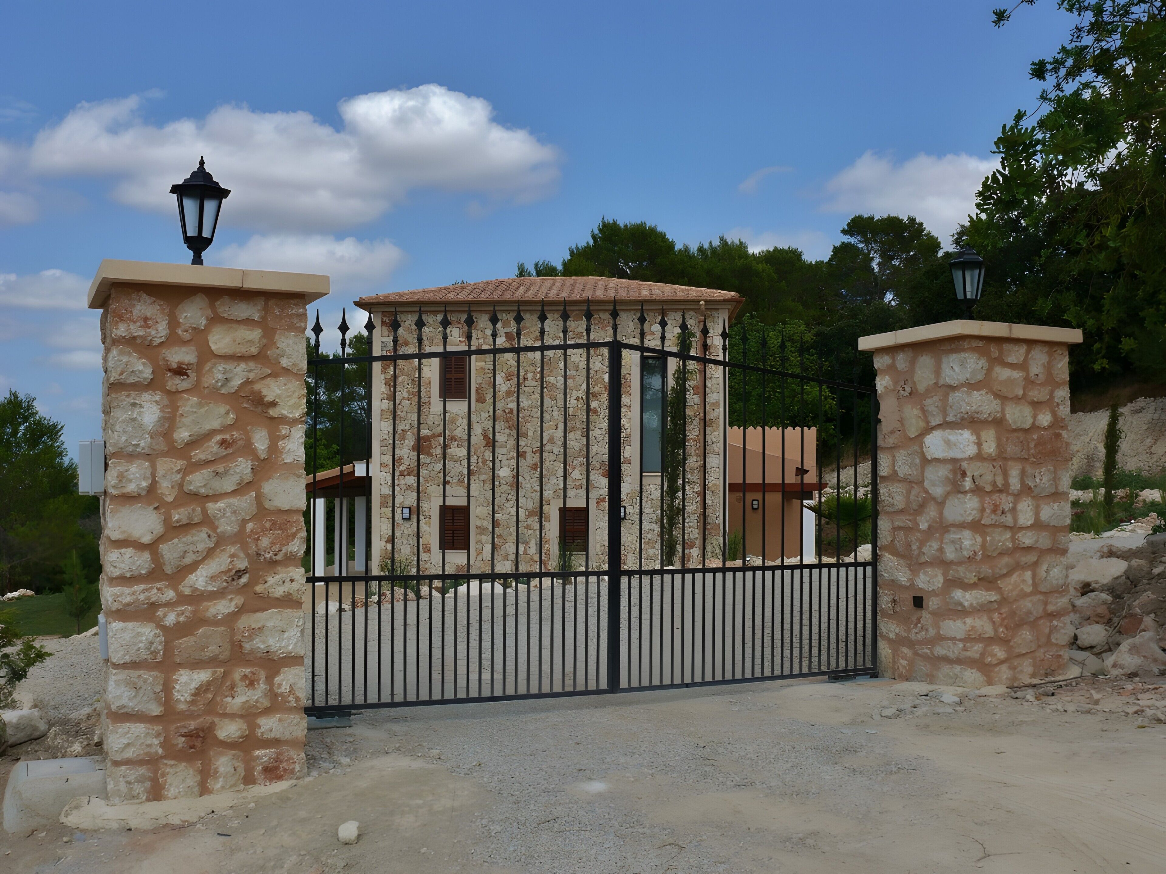 Une entrée fermée par un portail, avec des piliers en pierre et un portail en métal, qui mène à un bâtiment doté d’un toit en tuiles et de murs en pierre.