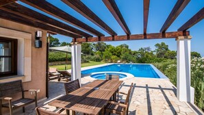 Ein überdachter Außenbereich mit Pool, Holzmöbeln und Blick auf die umliegende Grünfläche.