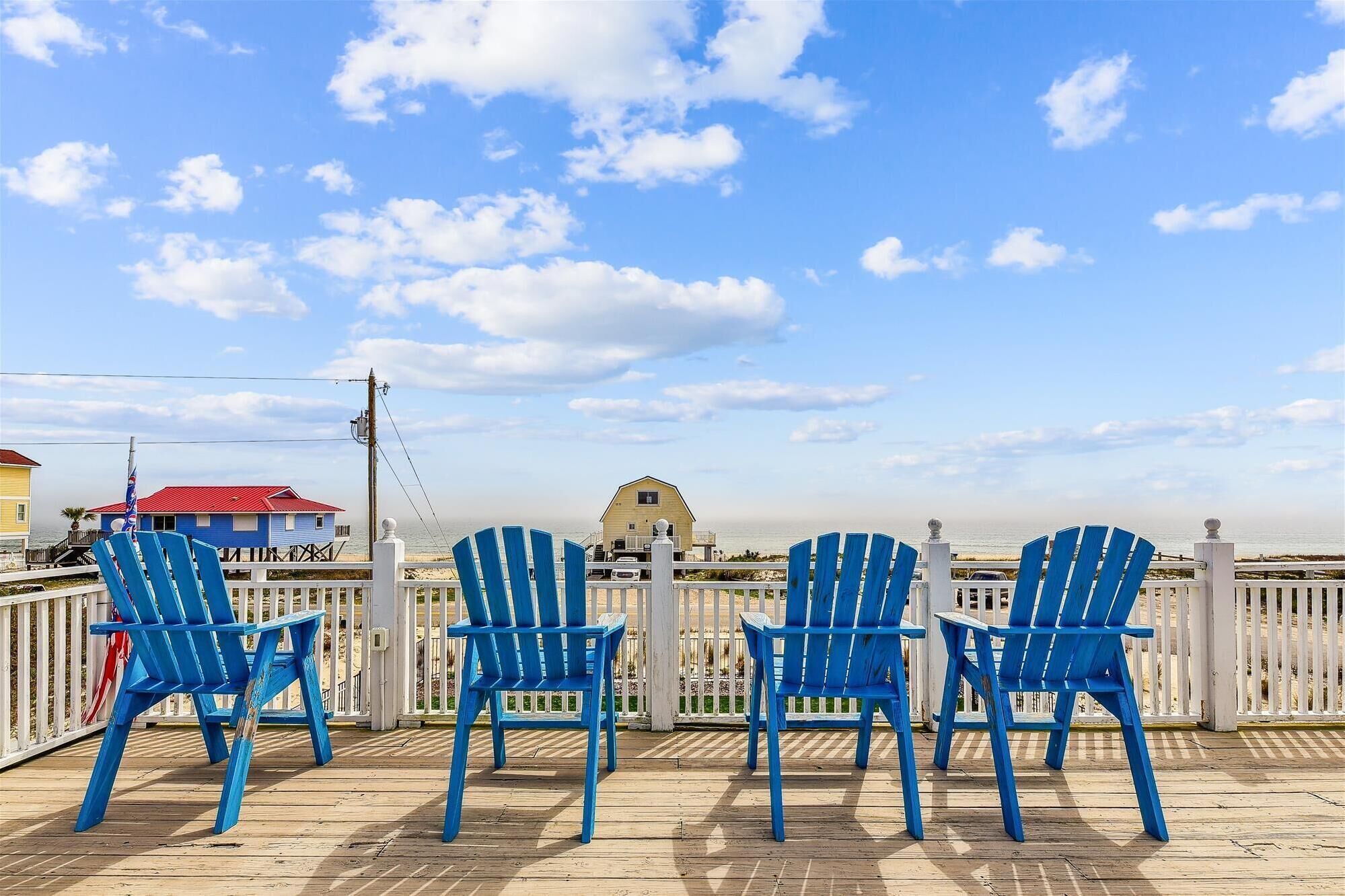 Eine Holzterrasse mit blauen Adirondack-Stühlen, die auf einen Strand ausgerichtet sind.