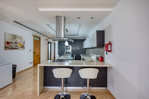 A modern kitchen with a central island, two bar stools, a stainless steel range hood, and a fire extinguisher mounted on the wall. - Modern and cosy 2 bedroom apartment PSCI1-1 (Mellieha)