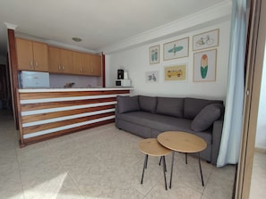 Ein Wohnzimmer mit grauem Sofa, zwei runden Holztischen, einer Küchenzeile mit Holzschränken und Wandbildern, die Surfbretter, Fahrräder und einen Van zeigen.