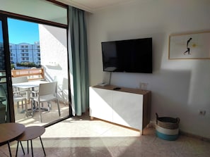 Ein modernes Wohnzimmer mit einem Flachbildfernseher, einem Essbereich mit Tisch und Stühlen, einer kleinen Kommode und einem großen Fenster mit Vorhängen.