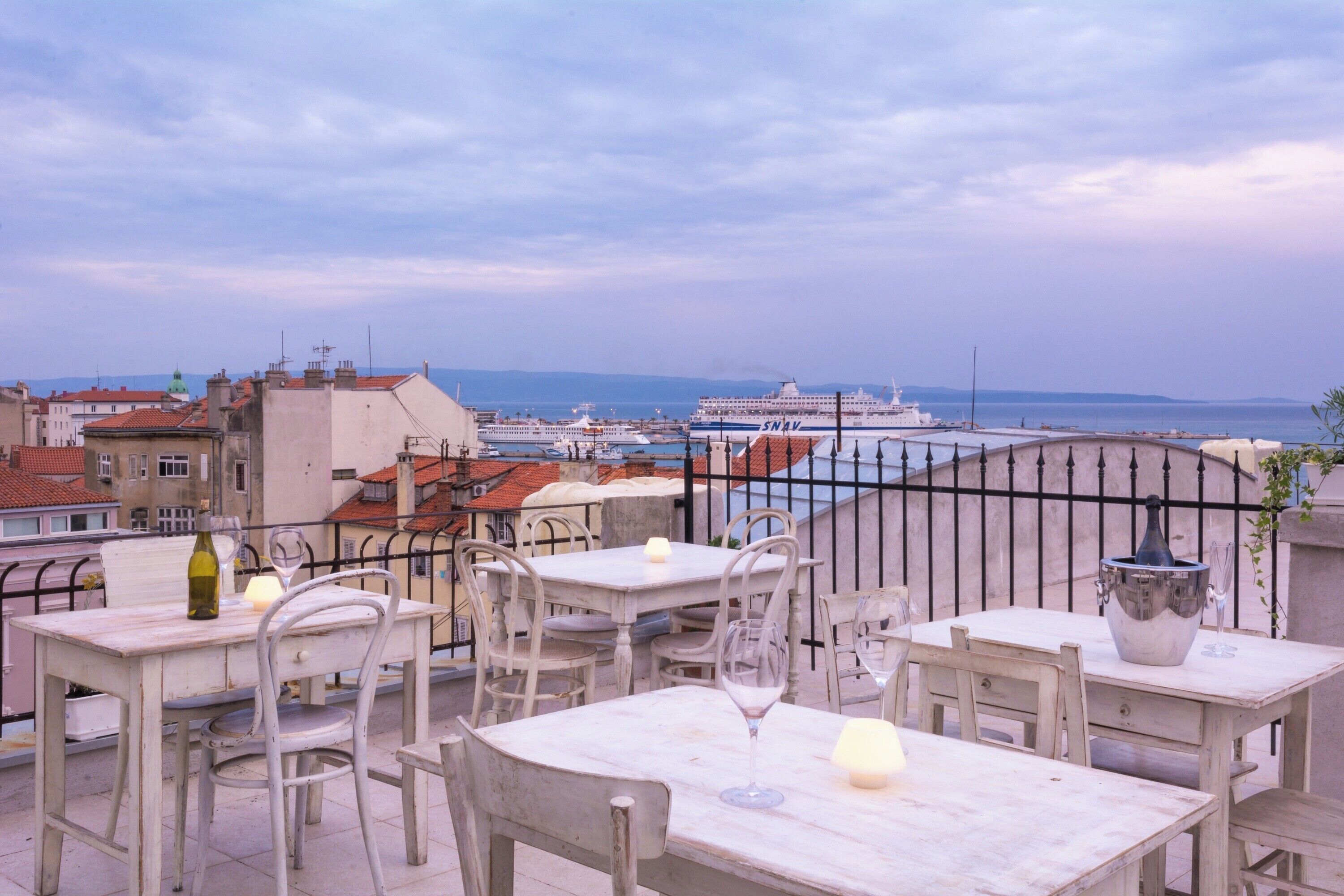 Eine Dachterrasse mit Tischen und Stühlen, mit Blick auf ein Gewässer und ein Kreuzfahrtschiff.