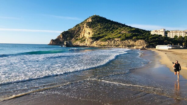 Una playa con una colina rocosa y edificios al fondo.