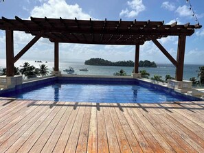 Een houten terras met een zwembad, een pergola en uitzicht op zee en een eiland.
