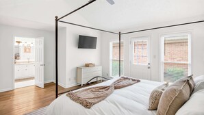 A bedroom with a four-poster bed, a ceiling fan, a television on the wall, and a view of a brick building through the windows. - Renovated 3BR Retreat With Game Room (Montgomery)
