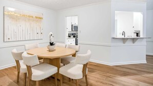 A dining area with a round wooden table and six upholstered chairs, a white wall with a large abstract painting, and a kitchen visible through an open doorway. - Renovated 3BR Retreat With Game Room (Montgomery)