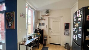 Uma cozinha compacta com um laptop sobre uma mesa, uma geladeira com ímãs, uma porta com uma pequena prateleira e uma janela com vista.