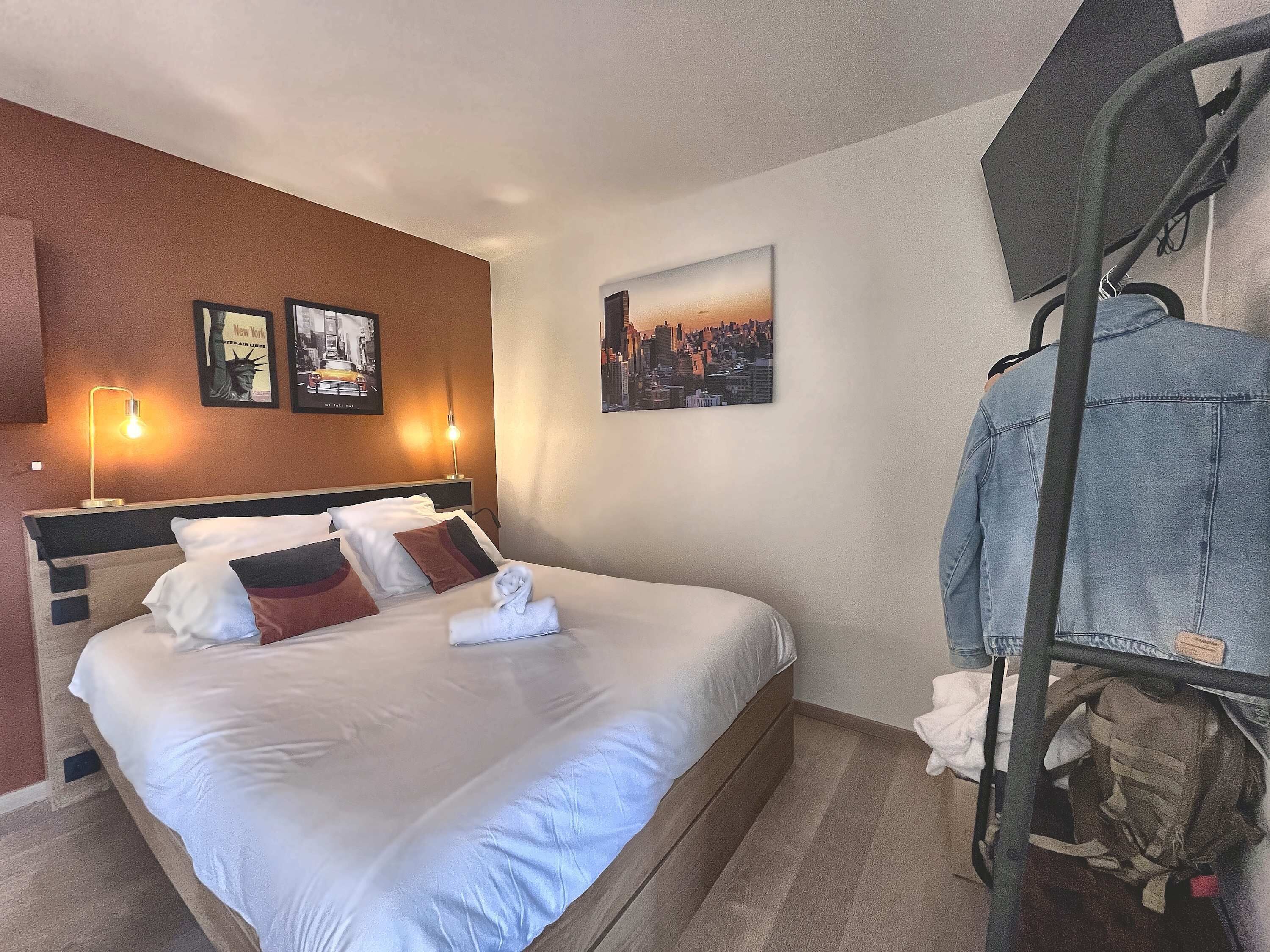 A hotel room with a bed, a headboard, a wall-mounted lamp, and a cityscape painting.
