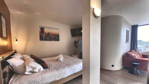 A hotel room with a bed, a cityscape painting, a TV, a chair, and a window. - The Manhattan - 2p- Over Toulouse s Roofs (Toulouse)
