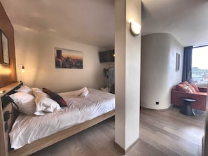 A hotel room with a bed, a cityscape painting, a TV, a chair, and a window. - The Manhattan - 2p- Over Toulouse s Roofs (Toulouse)