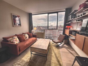 A modern living room with a sofa, a wooden coffee table, a chair, and a view of a cityscape through large windows. - The Manhattan - 2p- Over Toulouse s Roofs (Toulouse)