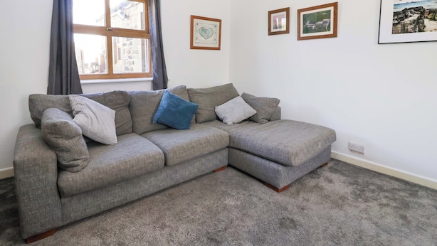 A grey sectional sofa with cushions, a window with curtains, and framed pictures on the wall. - 1 Upper Austby Farm Cottage (Ilkley)