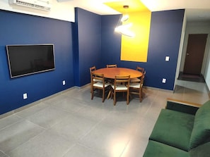 Quarto com parede azul, televisão de tela plana presa a ela, mesa redonda de madeira com cadeiras, sofá verde e luminária no teto.