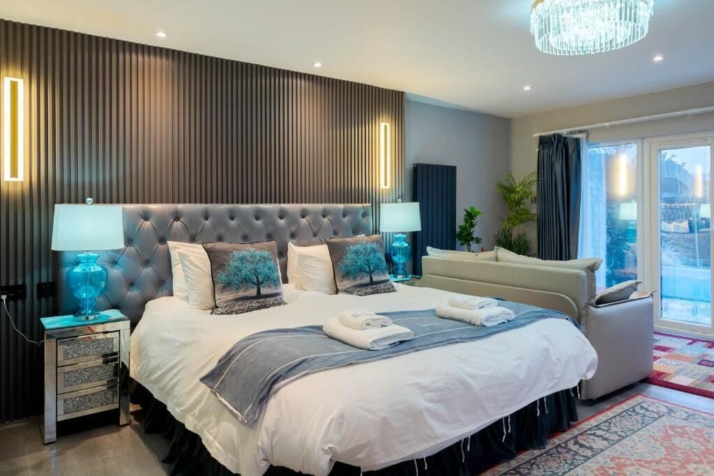 A modern bedroom with a tufted headboard, bedside lamps, a large bed with a grey and white duvet, a small side table, and a view through a sliding glass door.