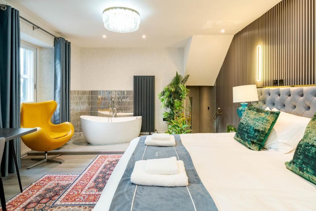Quarto de hotel moderno com uma cama grande, uma banheira, uma cadeira amarela e um candelabro.