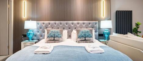 A modern hotel room with a tufted headboard, two bedside lamps, a large bed with a blue bedspread, and a vertical wooden panel wall.