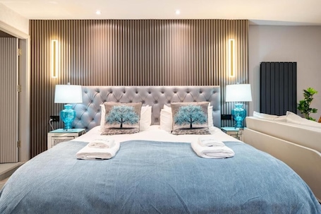 A modern hotel room with a tufted headboard, two bedside lamps, a large bed with a blue bedspread, and a vertical wooden panel wall.