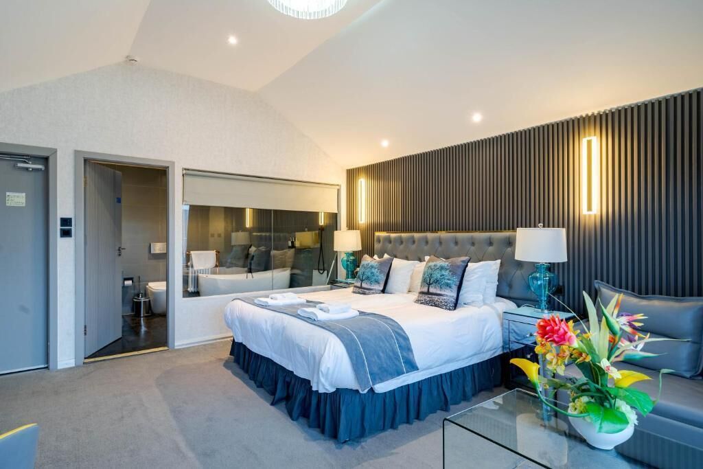 Un dormitorio moderno con una cama grande, una mesa de centro de cristal, un sofá, un espejo y una lámpara fijada a la pared.