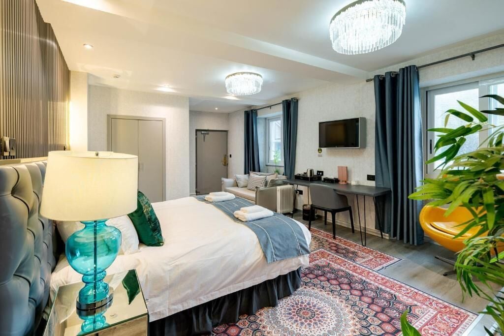 Quarto de hotel com cama, escrivaninha, cadeira, televisão e uma janela grande com cortinas.
