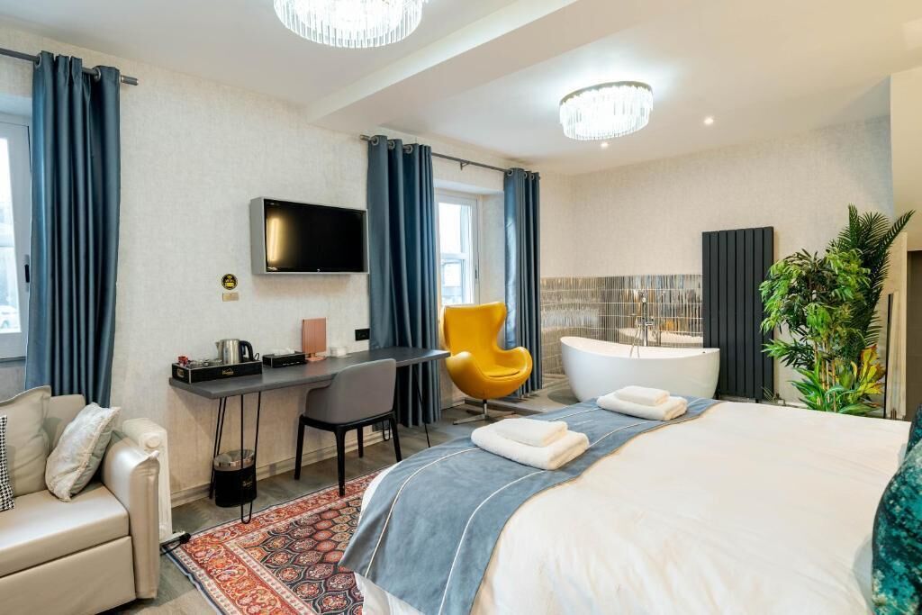 A hotel room with a bed, a desk, a chair, a television, a bathtub, and a potted plant.