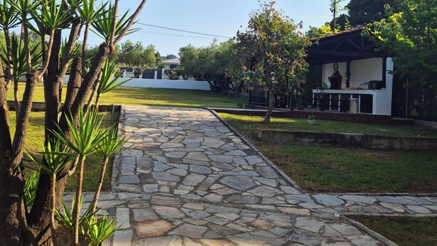A stone pathway leading to a building with a white exterior and a covered area with a statue. - GeoBasement - Mexico (Pefkochori)