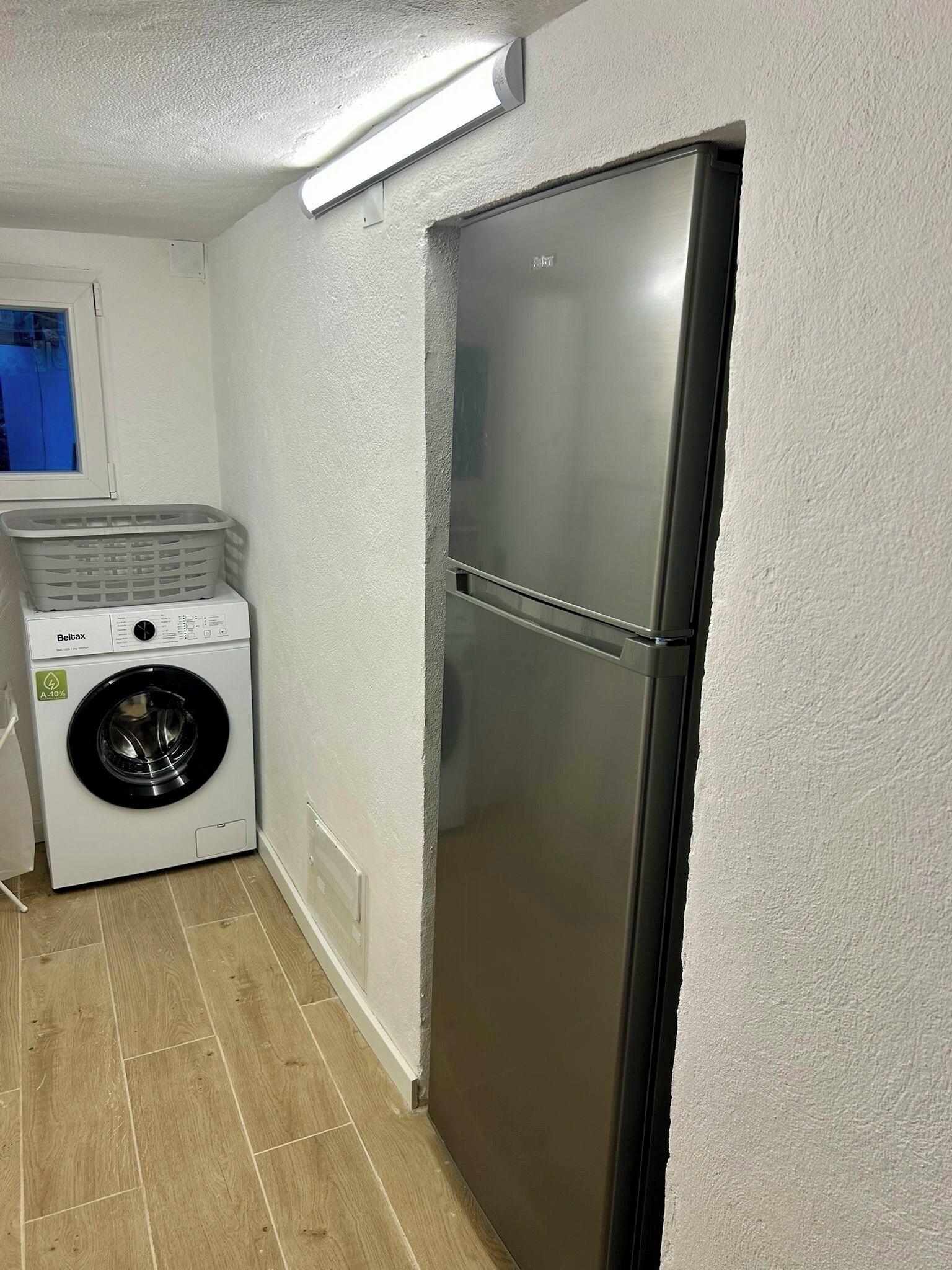 Un lavadero con lavadora de carga frontal, refrigerador de acero inoxidable y ventana con vista al exterior.