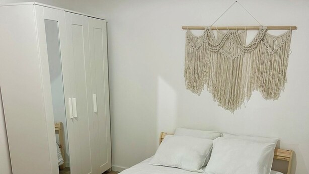 A neatly made bed with white linens, a macrame wall hanging, and a wooden wardrobe.