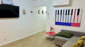 A modern living room with a flat-screen TV, a decorative wall piece, a round side table with a plant, a grey sofa with a green throw, and a colorful wall art piece.