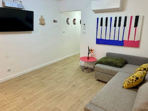 Un salón con un televisor de pantalla plana, una mesa redonda roja y negra, un sofá gris con una manta verde y un cojín amarillo, y una obra de arte con temática de piano y colores vivos en la pared.