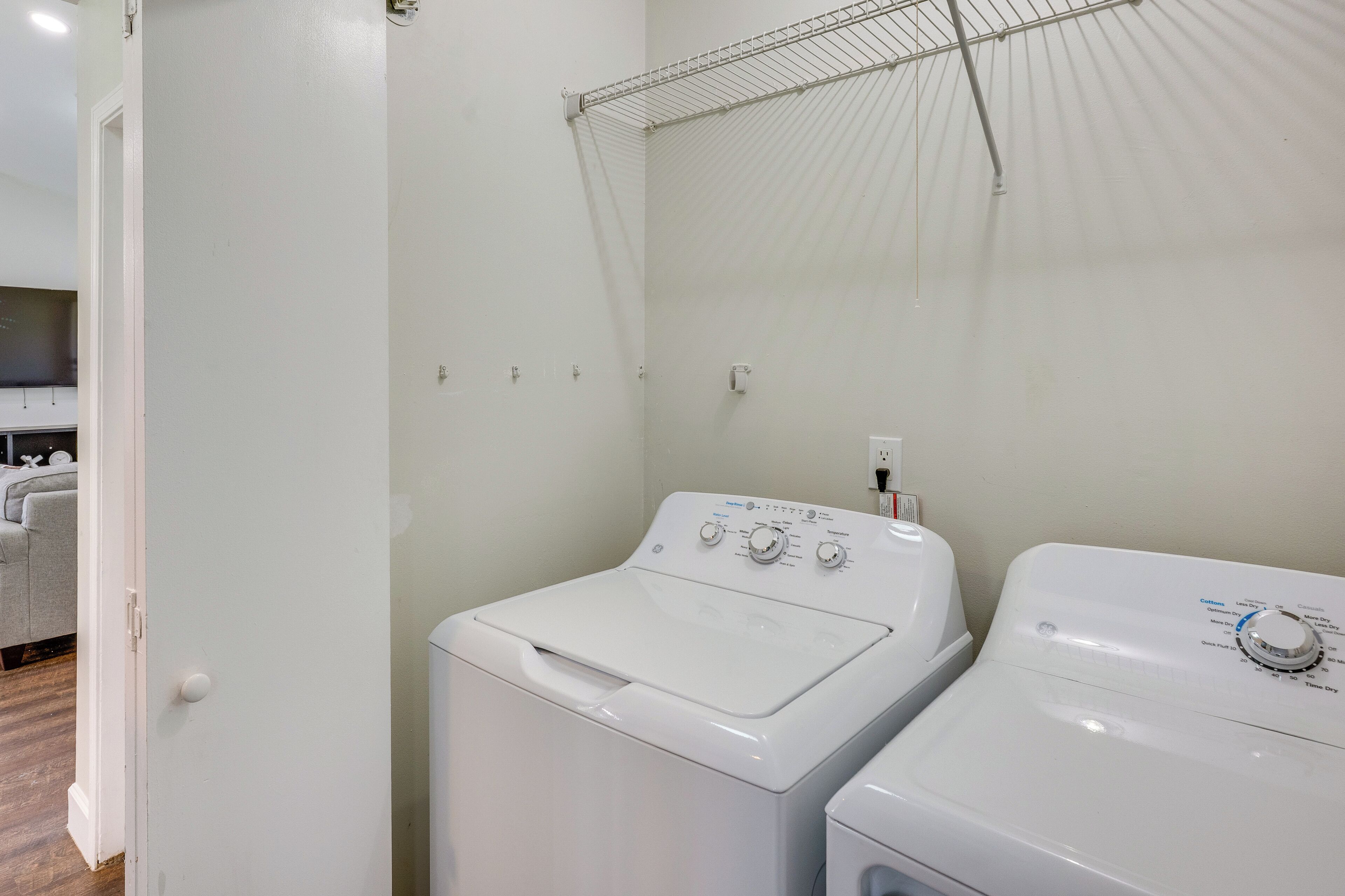 A laundry room with two white washing machines, a drying rack, and a door leading to a living area with a sofa.