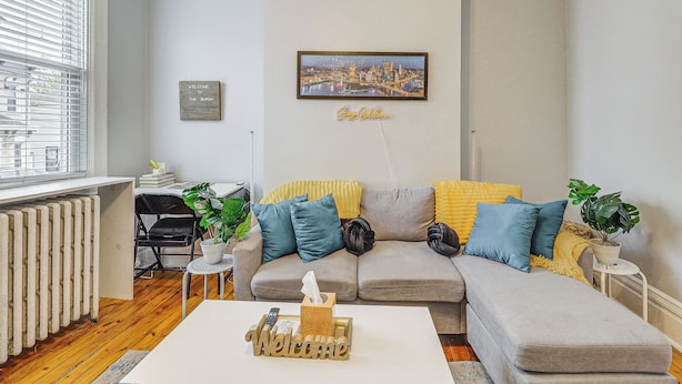 Een woonkamer met een bank, een salontafel, een radiator, een raam met jaloezieën, een stadsgezicht in een lijst en een welkomstbord.