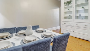 Een eetruimte met een tafel voor vier personen, blauwe gestoffeerde stoelen, een witte vaas en een wit kastje met glazen deuren.