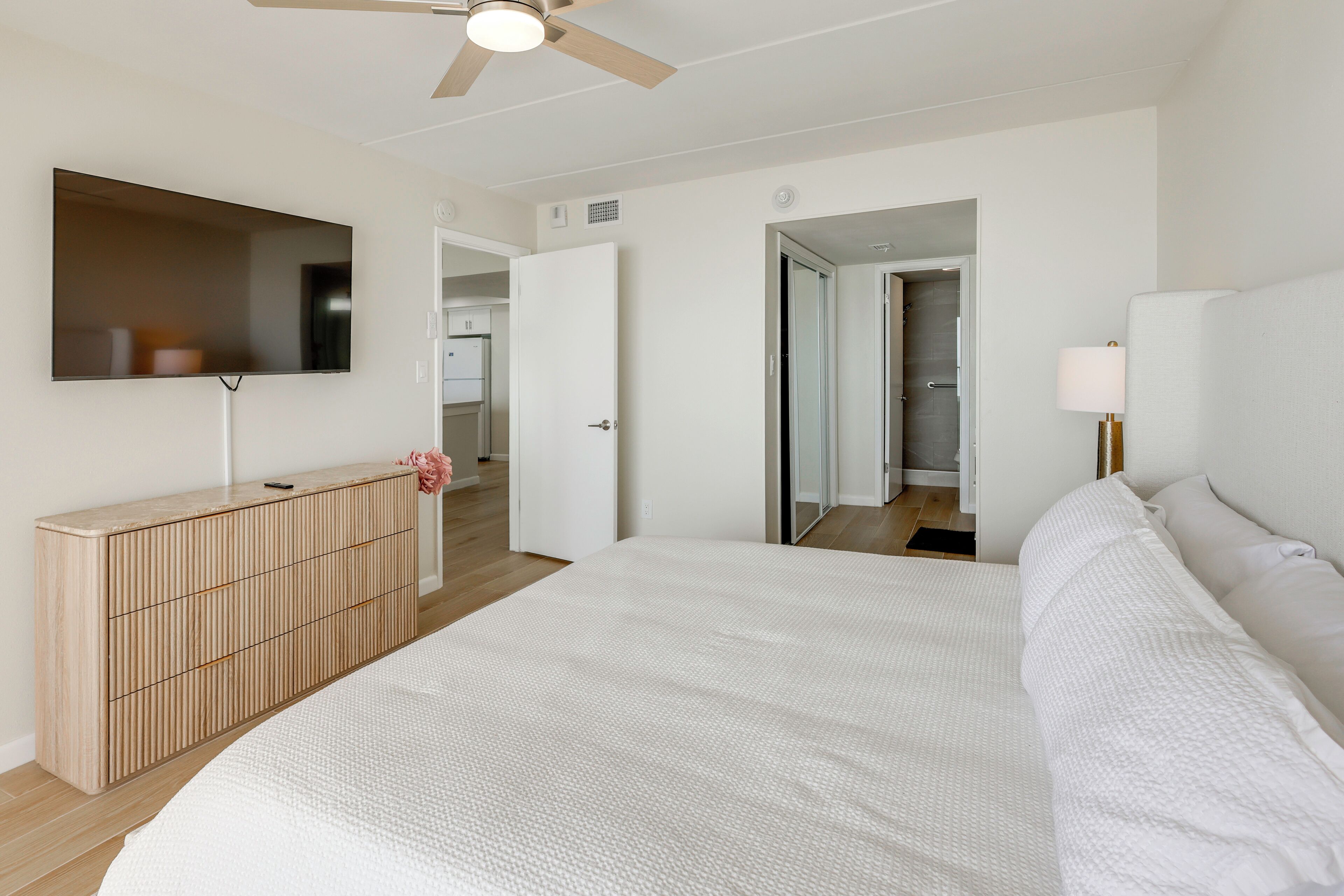 A bedroom with a bed, a dresser, a mirror, a ceiling fan, and a door leading to another room.