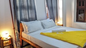 A bed with white bedding, a yellow blanket, and two pillows. There is a wooden headboard, a nightstand with a lamp, and a window with curtains. - The Hub Train House Exotic Roots (Zanzibar Town)