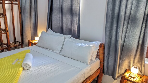A neatly made bed with white linens, a rolled-up towel, and a yellow blanket. There are two bedside lamps on wooden nightstands. - The Hub Train House Exotic Roots (Zanzibar Town)