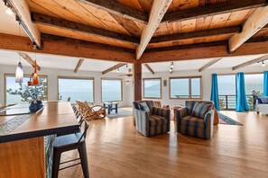 Une piĂšce spacieuse avec un plafond haut, des poutres en bois, un coin repas avec des chaises, un canapĂ© et de grandes fenĂȘtres offrant une vue sur la mer.
