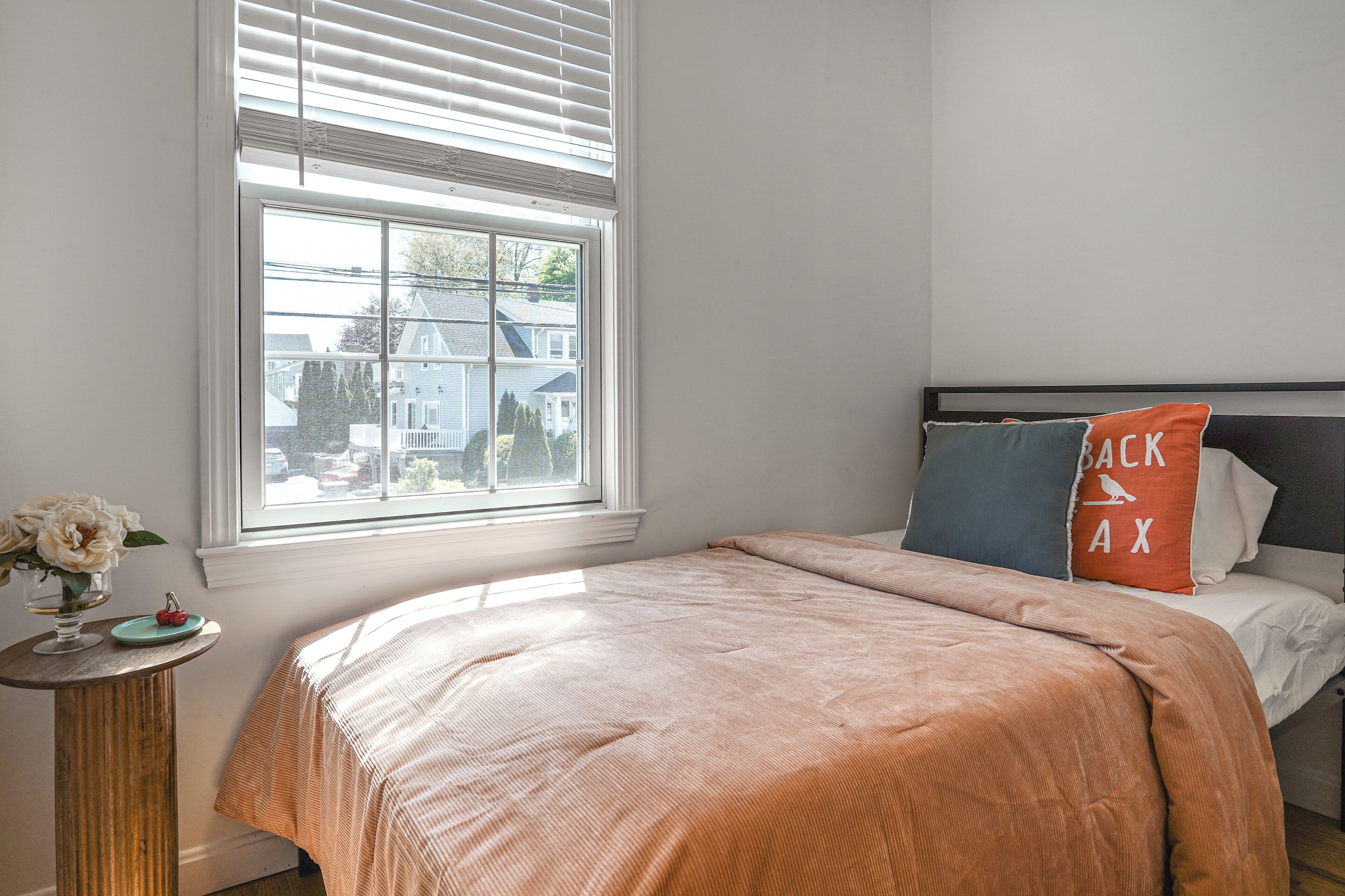 A bedroom with a bed, a window with blinds, a small table with a vase and a plate, and a view of houses outside.