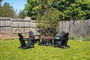 Un patio trasero con cerco de madera, una fogata y tres sillas Adirondack.