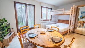 Una habitación con literas, una mesa de comedor con sillas, una planta en maceta y una ventana con cortinas.
