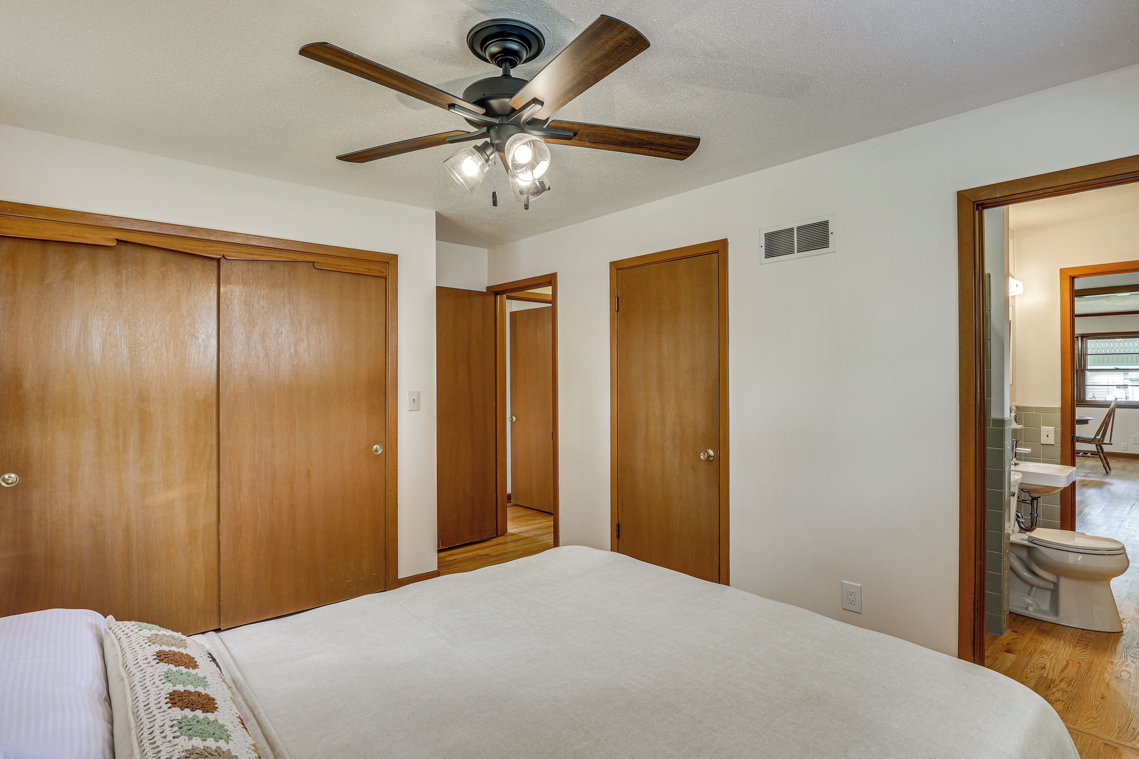 A bedroom with a bed, a ceiling fan, wooden wardrobes, and a bathroom visible through an open door.