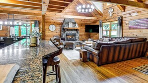 A log cabin interior with a stone fireplace, wooden ceiling beams, a bar area, and a leather sofa. - Hot Tub & Patio: Bedford Log Cabin on 10 Acres! (Moneta)
