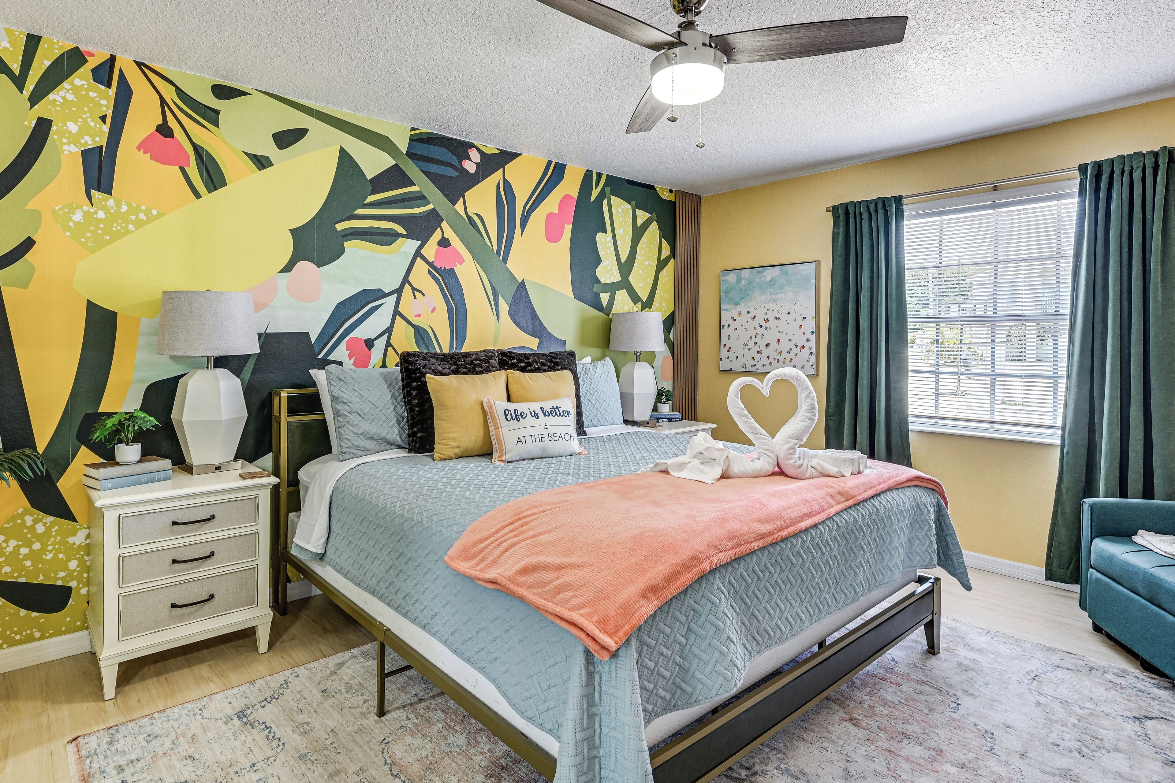 A bedroom with a bed, nightstands, a ceiling fan, a window with curtains, and a colorful wall mural.
