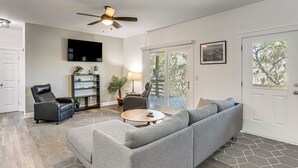 A living room with a gray sectional sofa, a round coffee table, a leather chair, a TV mounted on the wall, and a ceiling fan. - < 1 Mi to Ocean! OBX Condo w/ Decks, Pets Welcome (Hatteras)